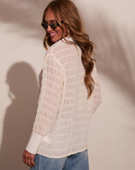 Woman wearing cream textured grid-pattern long-sleeve blouse with white cuffs, paired with blue jeans and round sunglasses