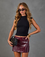 Woman wearing black crop top and burgundy vinyl mini skirt, holding black woven leather clutch, with gold accessories and cat-eye sunglasses
