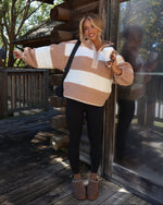 A woman stands on a rustic wooden deck in front of a log cabin, wearing a fuzzy quarter-zip pullover sweater with horizontal beige and off-white stripes, black leggings, and brown slippers.
