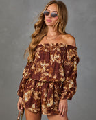 A woman modeling an off-the-shoulder floral romper with long puff sleeves and matching shorts in brown and beige tones, paired with round sunglasses, against a textured gray studio background.