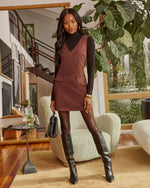 A woman models a brown suede-look sleeveless mini dress over a black long-sleeve turtleneck, styled with black tights, black knee-high boots, and a black handbag in a modern living room setting.