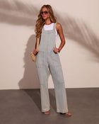 Woman wearing light wash wide-leg denim overalls over white tank top, styled with aviator sunglasses and woven crossbody bag