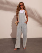 Woman wearing light wash wide-leg denim overalls over white tank top, styled with sunglasses, hoop earrings, and woven bag