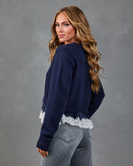 Woman wearing navy blue cropped sweater with white lace hem trim and gray jeans, posed from behind looking over shoulder