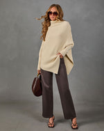 A woman models a cream ribbed knit poncho sweater, paired with dark brown wide-leg trousers, black strappy sandals, and a brown leather handbag, set against a textured gray studio backdrop.
