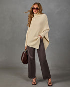 A woman models a cream ribbed knit poncho sweater, paired with dark brown wide-leg trousers, black strappy sandals, and a brown leather handbag, set against a textured gray studio backdrop.
