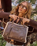A woman outdoors wearing oversized reflective sunglasses, a brown knit sweater, and holding a brown leather handbag with a white shearling-style lining and gold-tone hardware.