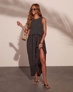 Woman wearing dark charcoal knit crop top over maxi dress with side slit, woven bag, gold jewelry, and black sandals