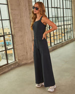 Woman wearing black wide-leg jumpsuit with square neckline, patch pockets, and silver sneakers in industrial setting