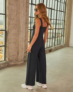 Woman wearing black wide-leg sleeveless jumpsuit with white chunky sneakers, posed in industrial loft setting