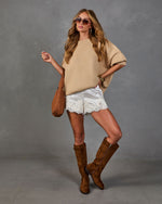 A woman models a beige knit poncho sweater, white lace-trimmed shorts, brown suede knee-high boots, and a brown shoulder bag against a textured gray studio backdrop.