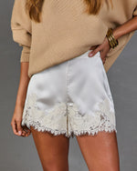 White satin shorts with a floral lace hem, worn by a model in a beige sweater against a gray studio background.