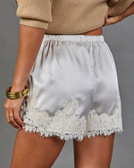 White satin shorts with a white floral lace hem, modeled with a beige ribbed knit top against a solid gray background.