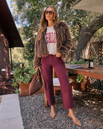 A woman stands outdoors on a patio wearing a brown faux fur jacket, burgundy straight-leg jeans, a cream graphic t-shirt, and beige pointed-toe flats. She is holding a brown woven handbag.