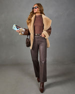 A woman models a brown faux-shearling jacket with dark brown leather sleeves, paired with a brown top, dark brown leather pants, and brown ankle boots, against a gray studio background.