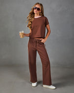 Woman wearing brown crop tee with pink collar and wide-leg track pants with white side stripe, holding iced coffee, white sneakers