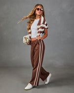 Woman wearing brown wide-leg track pants with cream stripes, striped top, white sneakers, and aviator sunglasses, carrying quilted clutch