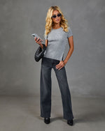 Blonde woman wearing gray ribbed short-sleeve top, dark wide-leg jeans, black heels, sunglasses, holding phone and woven clutch