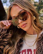 A close-up lifestyle shot of a woman wearing oversized aviator sunglasses with a red frame and dark brown lenses, styled with gold earrings and layered necklaces.