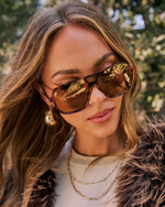 A close-up lifestyle shot of a woman wearing aviator-style sunglasses with dark brown frames and reflective gold lenses, set against a soft-focus outdoor background of green foliage.