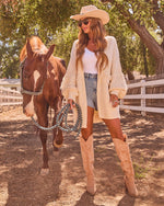 Woman in cream fringe cardigan, denim shorts, tan cowboy boots and hat, leading chestnut horse at sunny ranch