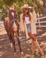 Woman in cowboy hat, fringe cardigan, denim shorts, and tan cowboy boots holding a chestnut horse at a ranch fence