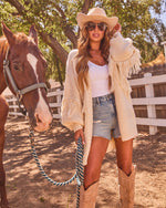 Woman in cream fringe cardigan, denim shorts, cowboy hat, and boots holding horse lead rope at ranch fence