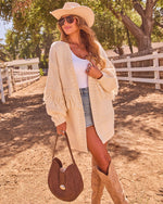 Woman in cream fringe cardigan, white tank, denim shorts, cowboy hat, tan boots, holding brown suede bag at sunny ranch
