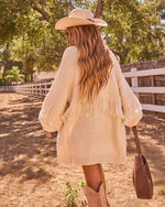 Woman in cream fringe knit cardigan, cowboy hat, tan boots, and brown tote walks along ranch fence path