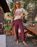 A woman stands outdoors on a patio wearing a cream sweatshirt with 'MERLOT' printed in burgundy across the chest, paired with burgundy straight-leg jeans and beige pointed flats. She holds a glass of red wine and carries a brown woven shoulder bag.