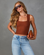 Woman wearing rust brown ribbed knit crop tank top, high-rise light wash jeans, aviator sunglasses, holding cognac leather clutch