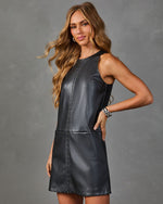 A woman modeling a sleeveless black faux-leather mini dress with decorative stitching and silver-tone studs, posed against a textured gray studio background.