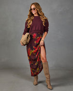 A woman models a two-piece outfit consisting of a crewneck long-sleeve sweater in deep burgundy and a matching high-slit midi skirt with a red and yellow floral print. She is styled with tan knee-high boots, sunglasses, and a tan clutch bag against a textured gray studio background.