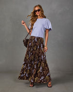 Woman in a lavender puff-sleeve top and brown floral pleated maxi skirt with sunglasses, crossbody bag and strappy heeled sandals