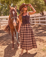 Woman in western cowboy hat, plaid midi skirt, and lace-up top leading chestnut horse at sunny ranch corral
