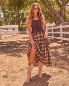 Woman in brown corset top, plaid maxi skirt, tan cowboy boots, and sunglasses posing on rustic ranch dirt path