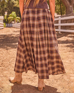Woman in plaid maxi skirt and tan cowboy boots walking dirt path by white ranch fence, back view