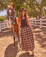 Woman in western cowgirl outfit with plaid midi skirt, brown corset top, and cowboy hat leading chestnut horse at ranch