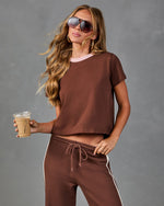 Woman in brown cropped tee and matching side-stripe sweatpants holding iced coffee, wearing aviator sunglasses