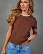 Woman wearing brown cropped ringer tee with pink collar and blue high-waist jeans, wavy blonde hair, hoop earrings