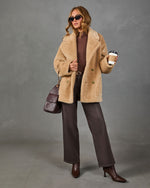 A woman models a beige shearling-style double-breasted coat over a dark brown turtleneck, paired with dark brown wide-leg trousers and brown ankle boots. She is holding a coffee cup and wearing a dark brown handbag against a textured gray studio background.