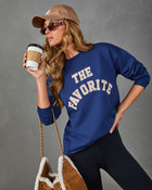 A woman wearing a navy blue sweatshirt with 'THE FAVORITE' printed in cream varsity lettering, paired with black leggings, a tan baseball cap, sunglasses, and a brown quilted handbag, holding a coffee cup against a gray studio background.