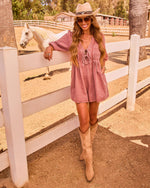 Woman in dusty rose romper, tan cowboy boots and hat poses by white ranch fence with white horse in background