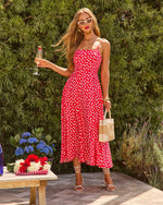 Woman in red floral midi dress with spaghetti straps holding champagne, wearing white strappy heels and carrying straw bucket bag outdoors