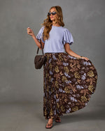 Woman in lavender puff-sleeve top and brown pleated floral maxi skirt with brown woven saddle bag and strappy sandals