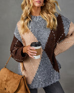 A woman wearing a chunky knit sweater with a large-scale geometric pattern in heather gray, dark brown, and beige. She is holding a brown coffee cup and wearing a brown suede shoulder bag, set against a solid gray studio background.