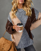 A woman wearing a chunky knit sweater with a large-scale geometric pattern in heather gray, dark brown, and beige. She is holding a brown coffee cup and wearing a brown suede shoulder bag, set against a solid gray studio background.
