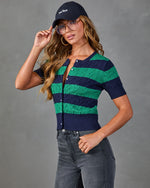Woman wearing navy and green striped cable-knit crop cardigan, gray jeans, New York baseball cap, and oversized glasses