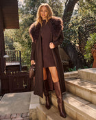 A woman stands on outdoor stone steps wearing a long dark brown coat with a shawl collar, a ribbed brown knit dress, knee-high brown boots, and holding a small woven handbag.