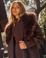 A woman wearing a dark brown knit sweater and a dark brown coat with a voluminous dark brown faux fur collar, standing outdoors in front of trees.